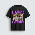 A black short-sleeve graphic t-shirt featuring five shocked cats with wide eyes and open mouths. Purple wavy text above the cats reads 'LESBIANS' and purple wavy text below reads 'EAT WHAT?!'.