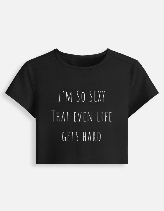 A black short-sleeved cropped baby tee featuring the text 'I'M SO SEXY THAT EVEN LIFE GETS HARD' in a thin, white, minimalist handwritten font.