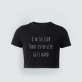 A black short-sleeved cropped baby tee featuring the text 'I'M SO SEXY THAT EVEN LIFE GETS HARD' in a thin, white, minimalist handwritten font.